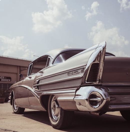 A vintage classic car with a sleek, chrome-detailed design is parked outdoors on a sunny day. The car has distinctive tail fins and a polished metallic finish, with a retro-style building visible in the background.